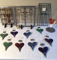Three stained glass window panes displayed standing on black stands, with ten colorful art glass hearts arranged in front on a white surface. One heart is mounted on a dedicated metal stand.
