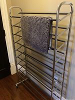Front and side view of a tall metal shoe rack with a grey plush rug draped over one of the horizontal bars, showing overall structure and texture.