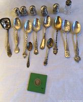 Overhead shot of all ten spoons, six sterling shakers, one glass shaker and packaged dime laid out on cloth.