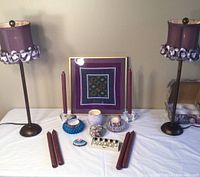 Photo showing two violet lamps with floral trimmed shades, framed quilt wall art, tall burgundy candles, glass candlestick holders, vintage bubble glass bowls, small candles and perfume bottles arranged on a table.