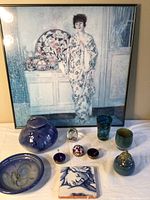 Overall lot showing the framed print, pottery, tile, art glassworks and paperweights arranged on white cloth.