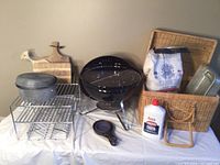 Photo of Weber charcoal grill, wicker picnic basket with contents including bag of charcoal, charcoal lighter fluid, cast iron pot, cooling racks, wooden cutting boards