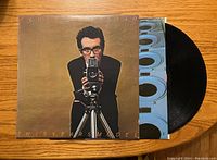 Front view of the vinyl record cover featuring Elvis Costello aiming a camera, with part of the black vinyl record visible inside.