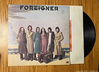 Front cover of Foreigner vinyl record showing the band members dressed in coats standing together with luggage by a beach, with the Foreigner logo at the top.