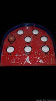 Close-up photo of red and blue metal skeeball game board featuring point-valued holes and one ball resting on surface.