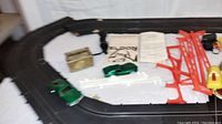 Main photo showing assembled black slot car track with two green slot cars, various red plastic supports, white guard rails, yellow electrical component, and instruction manuals on a white surface.