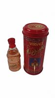 Photo showing the small glass bottle of Versace Red Jeans perfume and its red tin container with decorative gold and colorful elements, from an angled view highlighting the set.