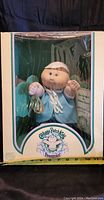 Front view of Cabbage Patch Kids Preemie Wilton Logan doll in original box. Doll dressed in light blue outfit with white trim, visible through clear plastic window.