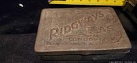 Closed view of rectangular metal tin with embossed Ridgways Teas London on lid and tape measure for scale.