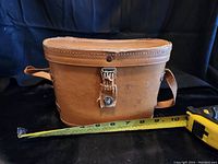 Leather carrying case measuring approximately 10 inches width with strap and snap closure shown alongside tape measure for scale