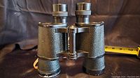 Black textured Kurt Muller binoculars with metal eyepieces placed on dark surface with tape measure