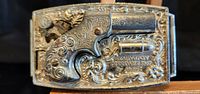 Close-up of the front side of the metal belt buckle designed as a revolver with floral pattern engravings.