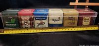 Photo showing six vintage-style tobacco tins in a clear plastic case, lined up with ruler for scale.
