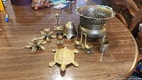 Photo showing the entire brass items set on wooden table: turtle figure, goose figure, bell, footed planter, three small cups, pair of candlesticks.