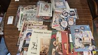 Collection of assorted leaflets, patterns, and cross stitch kits arranged on a table showing variety and themes including Hawaiian Islands and vintage toys.