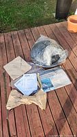 All firepit components laid out on wood deck: round metal firepit bowl, mesh spark screen wrapped in plastic, metal legs wrapped in paper, instruction manual, and other documentation.
