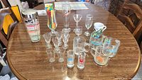 Wide shot of assorted glassware, cocktail shaker, and colorful cocktail stir sticks arranged on a wooden table.