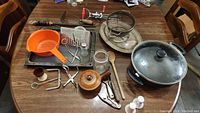 Overview of the kitchen items on a wooden table, showing frying pan with lid, metal roasting tray, orange colander, metal wire colander, wooden spoon, potato masher, egg beater, salt and pepper shakers, and other utensils.