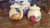 Set of three ceramic kitchen storage jars with fruit motifs on a wooden table.