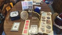 Photo showing assorted photo frames of different sizes and styles arranged on a wooden table. Includes oval gold-tone and various rectangular frames, some stacked.