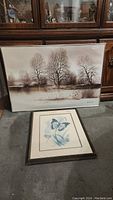 Two framed watercolour prints shown side by side on the floor: one of a butterfly and one of a landscape with trees and water.