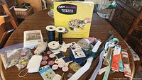 Overview of sewing accessories spread on a wooden table showing box, ribbons, threads, zippers, safety pins and other supplies