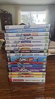 Photo showing stacked DVD game cases including Sims 3 base game and many Sims 3 and Sims 2 expansions, plus Sim City 3000 at bottom. Cases show titles clearly and verify disc presence.
