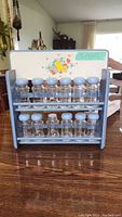 Front view of vintage blue and white wooden spice rack with a floral and butterfly design on the back panel. Two shelves hold 12 small clear glass spice jars with round blue lids.