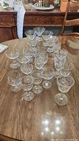 Photo showing a collection of 23 assorted stemmed glasses on a wooden table including multiple types of glasses such as brandy snifters, champagne, martini, and sherry glasses.