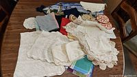 Overview of assorted table linens, doilies, scarves, and trivets arranged on wooden table showing details and variety of fabrics and designs.