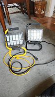 Two portable electric work lights on floor showing front and side views, one yellow and one black frame with metal cages and cords