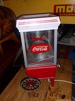 Front and side view of a vintage Coca-Cola branded popcorn machine display with glass panels, red and white metal body, and large wheel on one side.