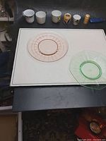 Photo of two small retro colored glass plates (pink circular with gold accents and green octagonal) and six small cups of various designs and colors on a table.