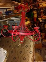Photo shows a red metal chandelier with five candle holder arms positioned on a beige patterned upholstered surface in a cluttered storage area.