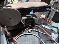 Side angle photo showing the metal belt sander with circular sanding surface and horizontal sanding belt. The device is plugged in with several visible cords and control knobs.