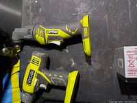 Two Ryobi cordless power tools placed on a flat surface, one reciprocating saw and one drill or driver, both yellow and black, no batteries attached.