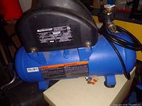 Front angled view showing brand label and warning stickers on blue cylindrical compressor tank with black motor housing.