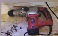Image of red and black corded hammer drill with metallic drill section placed on surface