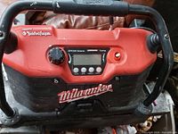 Front view of Milwaukee portable power station and radio showing control panel with volume, tuning knobs, and digital display. Black handles and red casing visible.