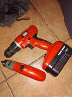 Photo shows two cordless power drivers on a tiled floor: one larger 18V cordless drill and one smaller 2.4V cordless screwdriver, both Black & Decker brand in orange and black colors.