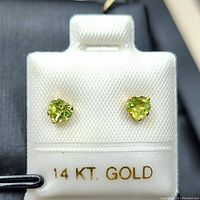 Close-up front view of the pair of heart-shaped natural peridot stud earrings in 14K gold on a white holder marked 14 KT GOLD.