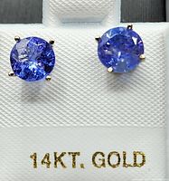 Two round bluish violet tanzanite stones mounted in four-prong white gold stud earrings displayed on white holder marked '14KT. GOLD'