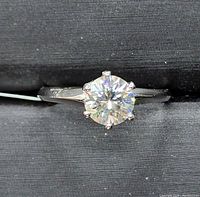 Close up image of a 10k white gold ring with a large round brilliant cut moissanite centerpiece mounted with six prongs.