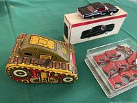 Overview photo of a 1930s tin tank toy with colorful graphics, 1963 black Ford Thunderbird diecast car on its original box, and a plastic container holding several small red metal cows, vintage red lead soldiers, and small metal trinkets.