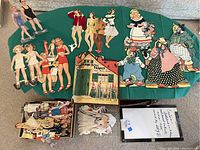 Full view of the paper doll collection including various paper dolls laid out on a green cloth, visible booklet or set labeled 'The Paper Doll Family', and piles of smaller paper doll accessories.
