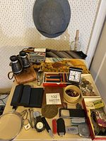 Wide view of all items including binoculars on leather case, neckties, wallets, medals, magnifying glasses, miniature collectible bottle, Timex Expedition wristwatch, collectible coin award and other small accessories on table with gray pith helmet above.