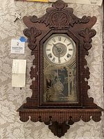 Front of the E. Ingraham gingerbread mantel clock showing carved wooden case, clock face with Roman numerals, and etched glass door with foliage and bird design. Winding key and paperwork nearby.