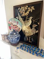 Full display of Asian themed artworks and collectibles including framed pheasant artwork, porcelain jar, decorative tin, fan, and blue beaded necklace.