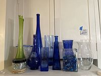 Overall view of assorted glass vases in blue, green, and clear colors with various shapes and a small container of decorative gems.