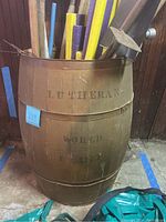 Front angled view of the large wooden barrel, showing faded stencil lettering 'LUTHERAN' and contents including foam pool noodles and tools.